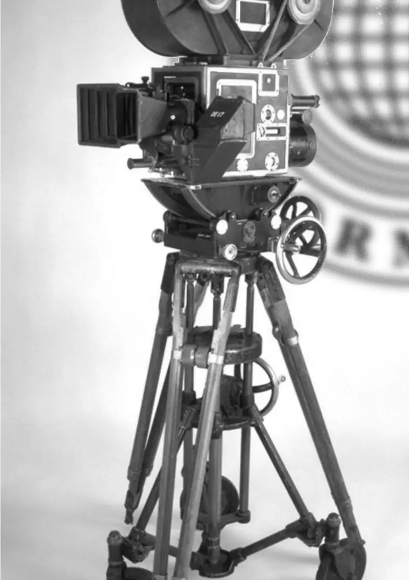 Black-and-white photograph of an antique film camera on a wheeled tripod, a blurred Department seal behind it.