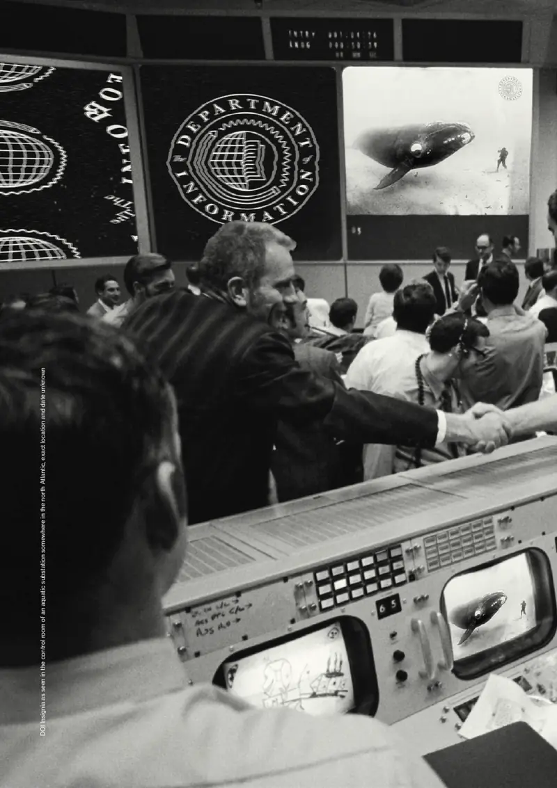 Black-and-white photograph of a crowded control room. The Department seal is projected on large screens above the consoles.