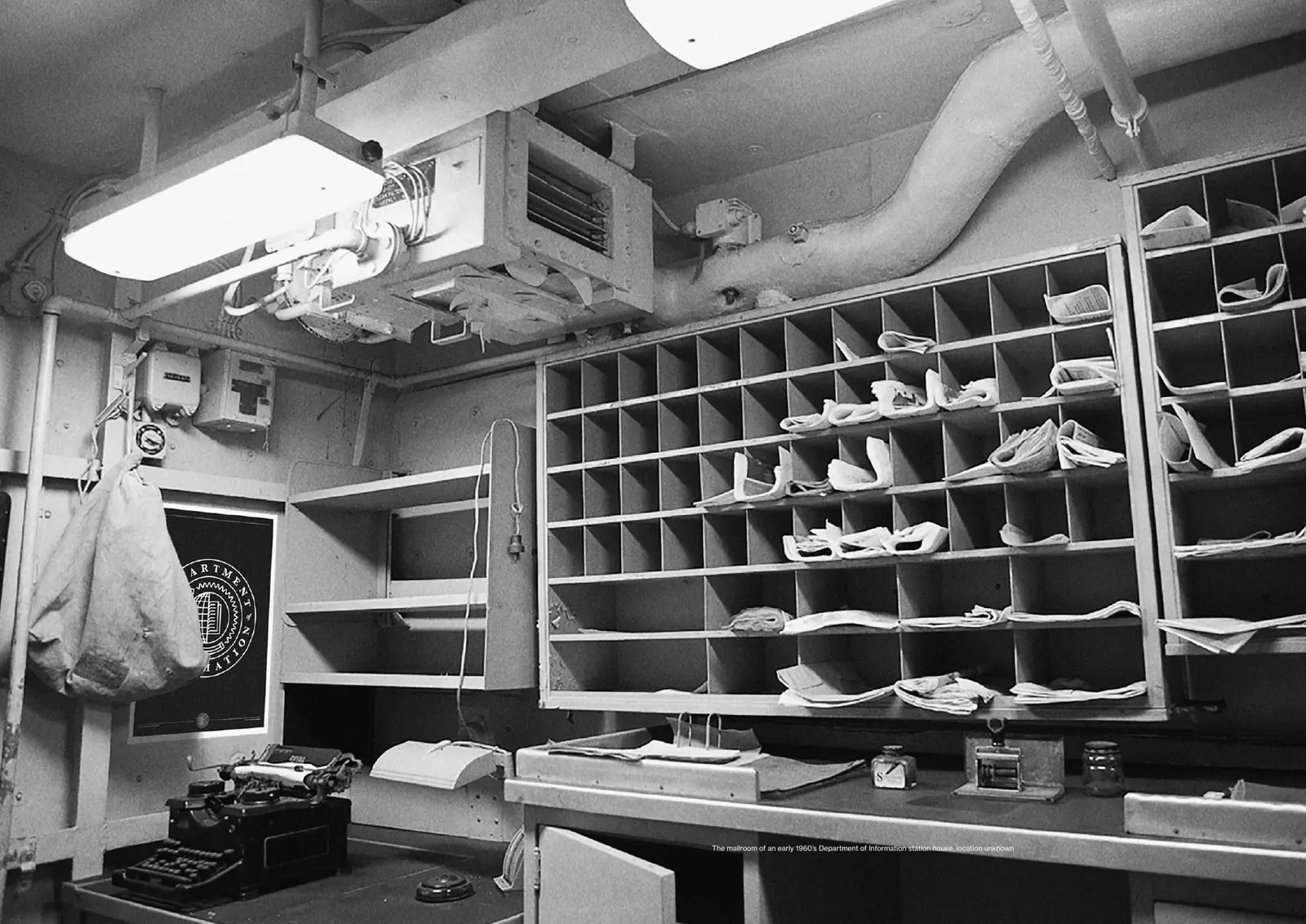 Black-and-white photograph of a cramped mailroom with pigeonholes full of papers, a typewriter and a framed Department poster.