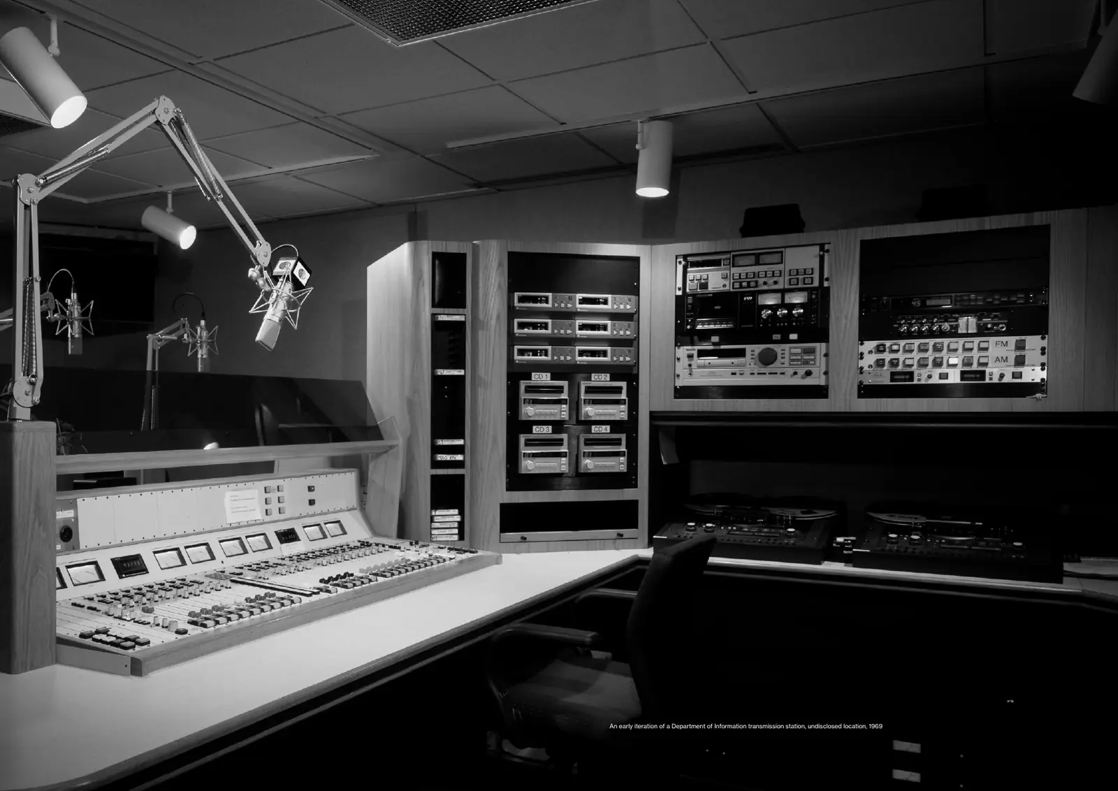 Black-and-white photograph of a radio studio with a mixing desk, microphones on boom arms and racks of tape and CD equipment.