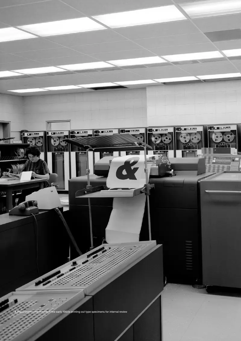 Black-and-white photograph of a 1960s computer room. A printer feeds out a large ampersand on continuous paper.